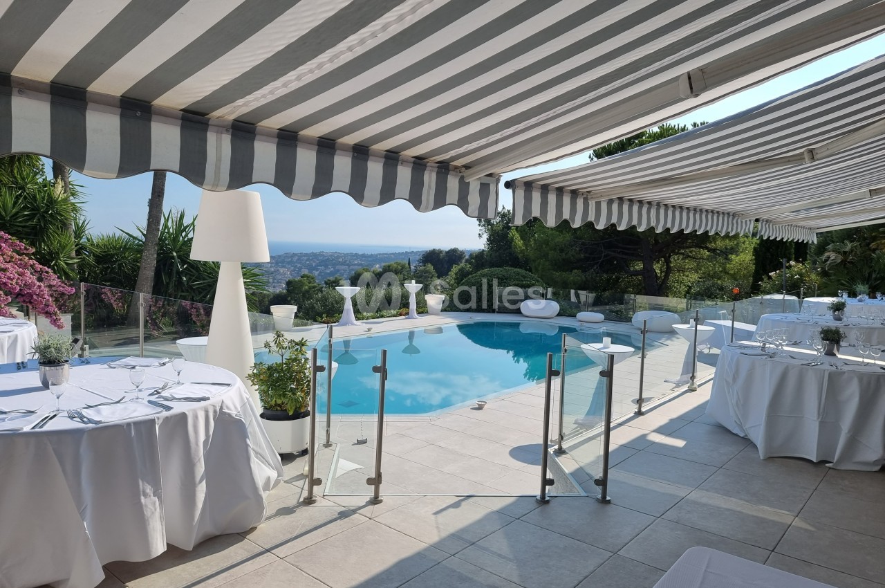 Terrasse avec piscine, tables dressées pour un repas, vue dégagée sur un paysage, ombragée par des stores rayés.