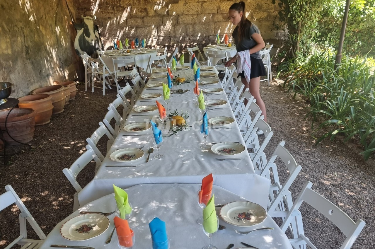 Tables longues dressées avec nappes blanches, assiettes et serviettes colorées, une personne ajuste les détails.