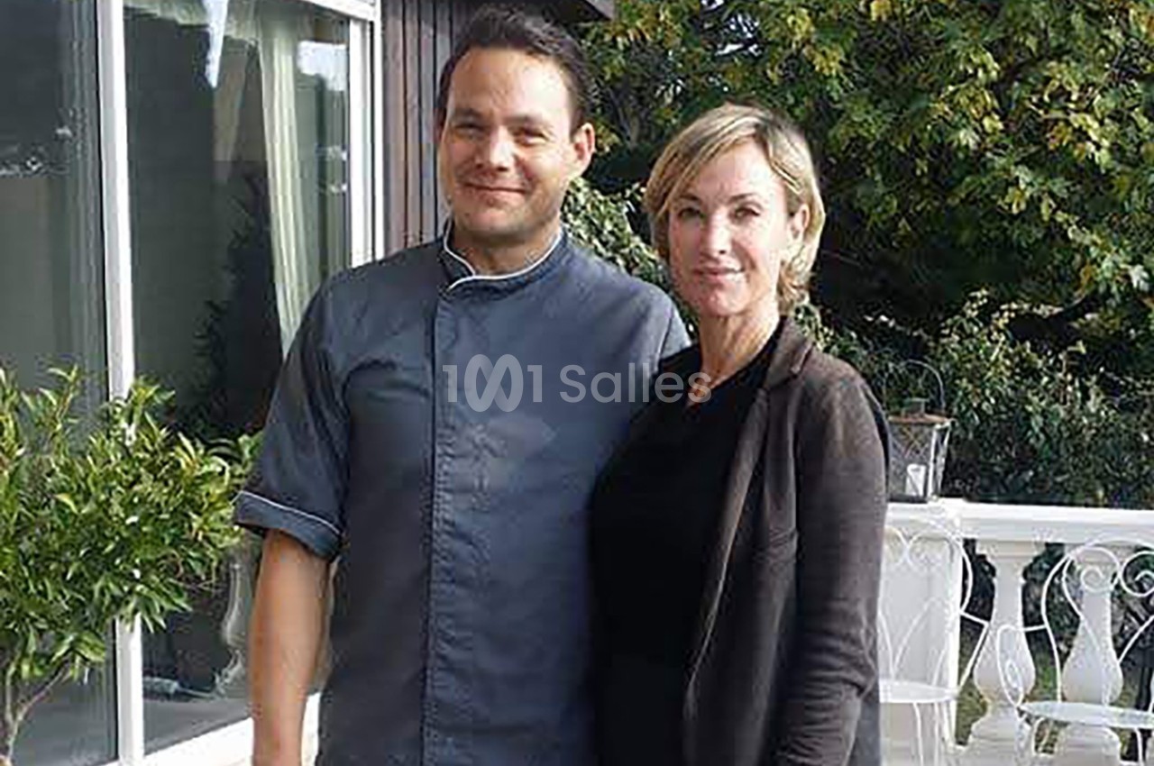 Un homme en tenue de chef et une femme posent ensemble sur une terrasse devant une maison et des arbres.