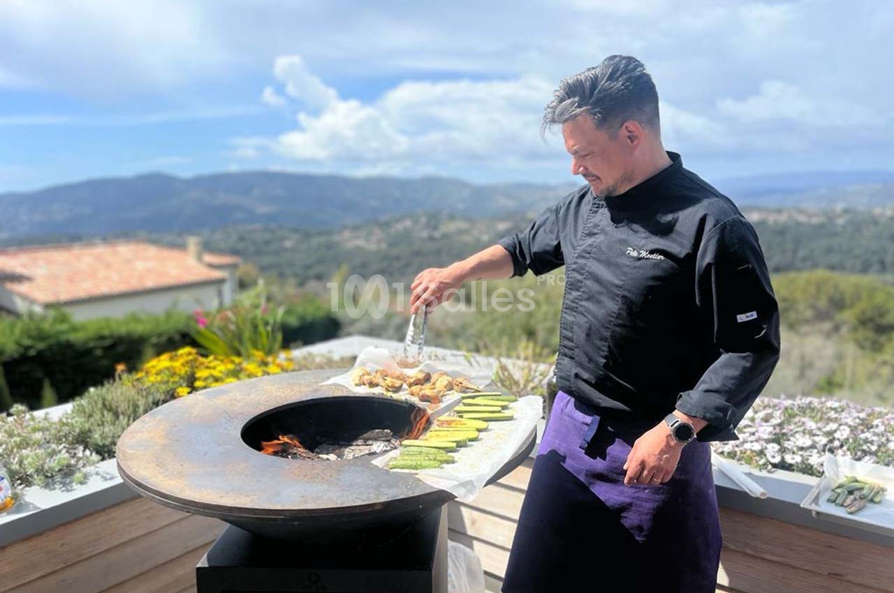Un chef cuisine des légumes et de la viande sur une plancha ronde en extérieur avec vue sur un paysage vallonné.
