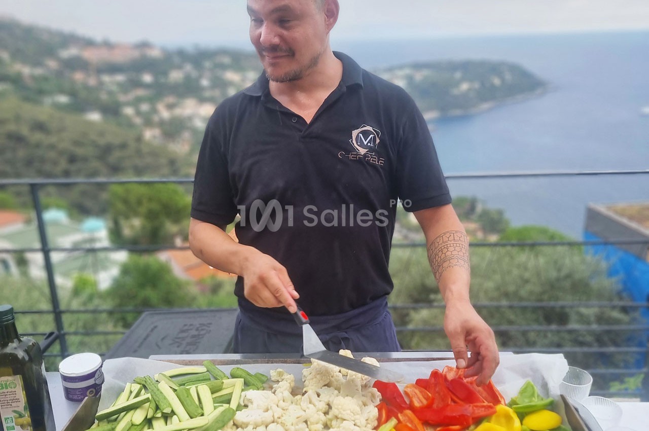 Un homme découpe des légumes frais sur une terrasse avec vue sur la mer en arrière-plan.