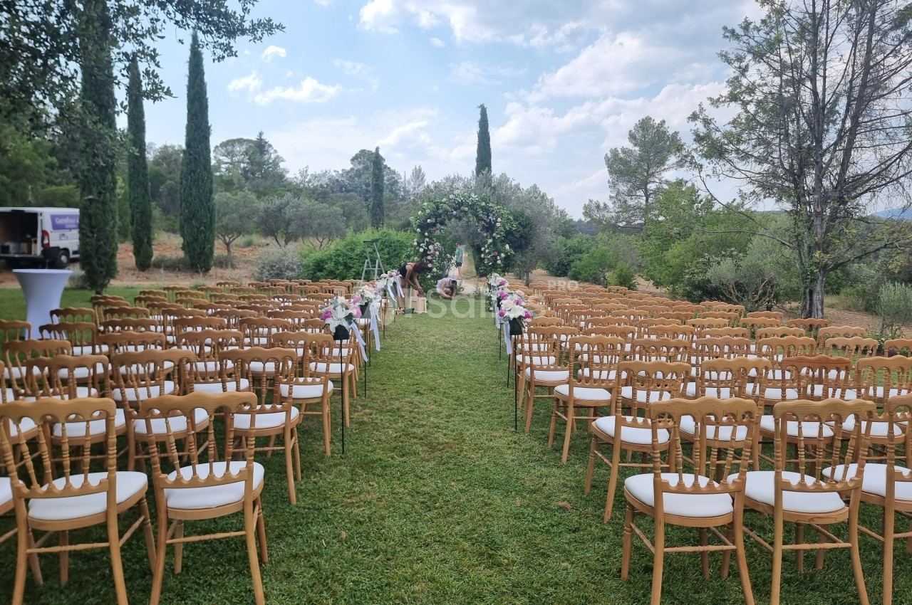 Allée centrale bordée de chaises en bois et décorée de fleurs, menant à une arche florale dans un jardin.