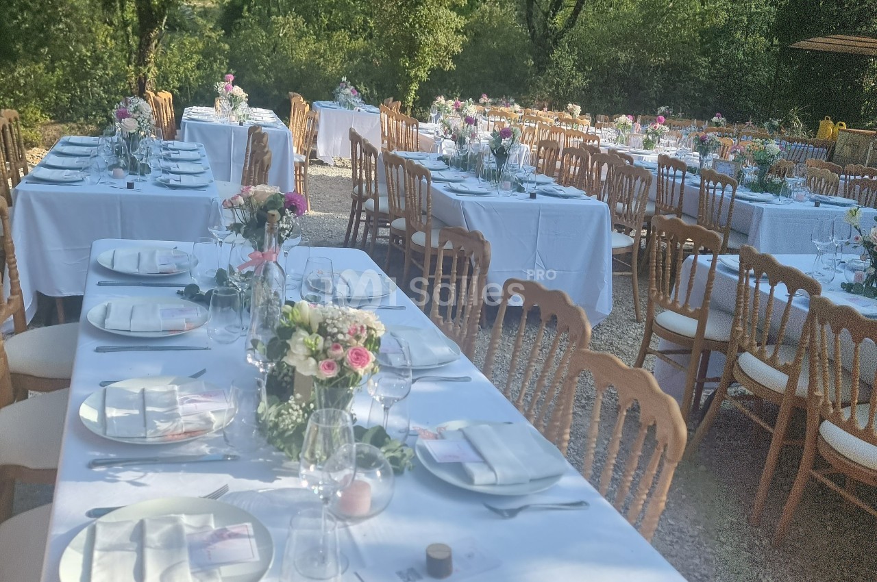 Tables de banquet dressées en extérieur avec nappes blanches, chaises en bois et décorations florales.