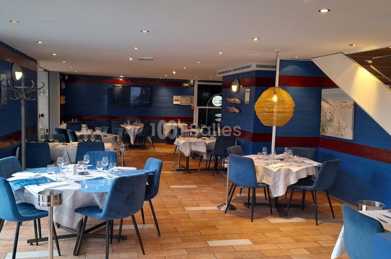 Salle de restaurant lumineuse avec tables dressées, chaises bleues, murs en bois bleu et décoration maritime.