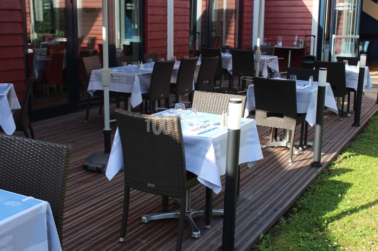 Terrasse d'un restaurant avec des tables dressées, chaises en osier et nappes blanches, sur un sol en bois.