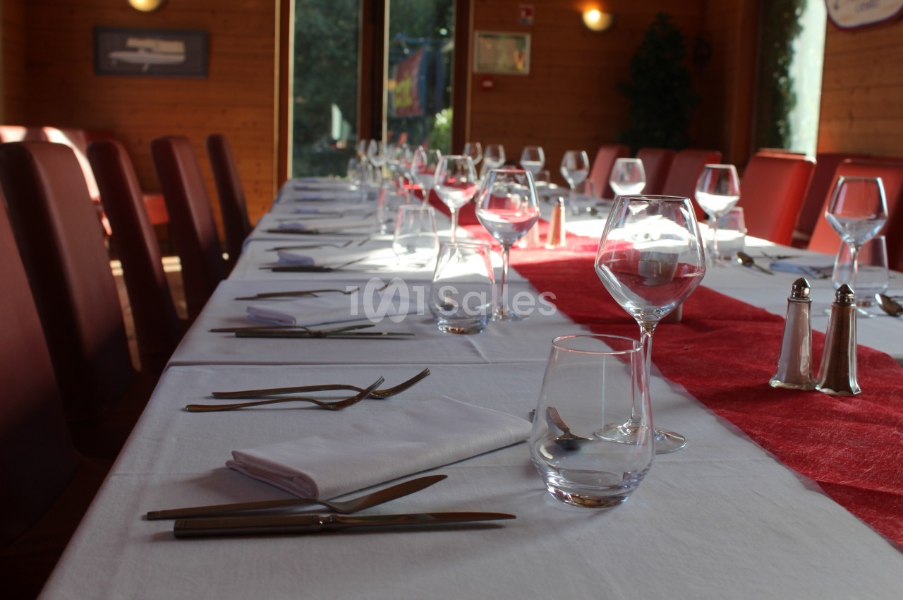 Table dressée avec nappes blanches, serviettes pliées, couverts, verres et chemin de table rouge dans une salle lumineuse.