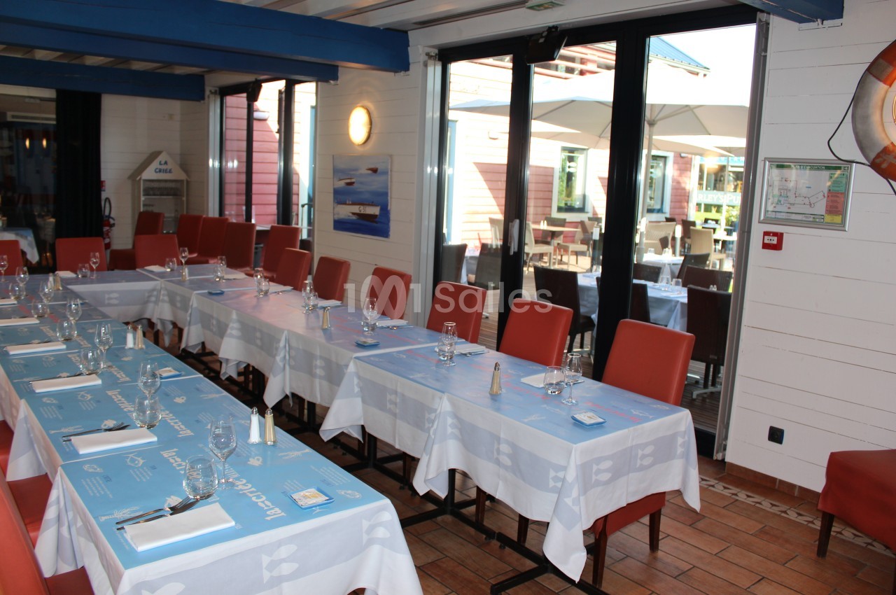 Salle de restaurant lumineuse avec tables dressées, nappes bleues et chaises rouges, donnant sur une terrasse extérieure.