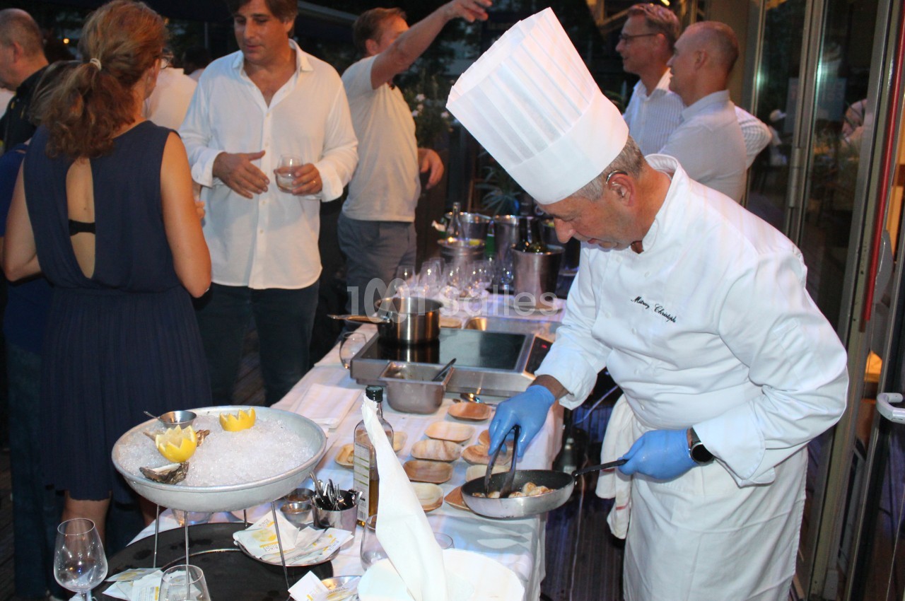 Un chef cuisinier prépare un plat lors d'un événement en plein air, entouré de convives discutant près d'une table dressée.