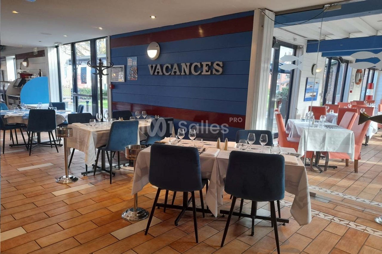 Salle de restaurant lumineuse avec tables dressées, chaises bleues et mur décoré du mot ’VACANCES’.