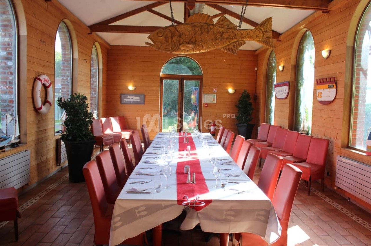 Salle de restaurant lumineuse avec une grande table dressée, entourée de chaises rouges et décorée d'éléments marins.