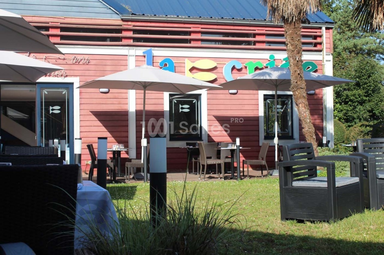 Terrasse d'un restaurant avec parasols, tables et chaises, devant une façade en bois rouge et un logo coloré.