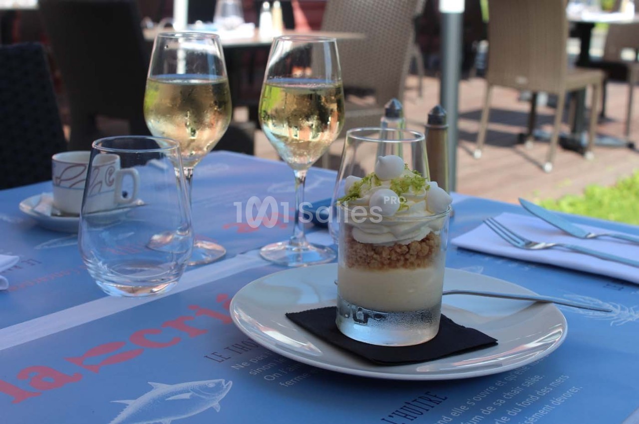 Dessert servi dans un verre avec mousse, crumble et zestes de citron, accompagné de deux verres de vin blanc sur une table…