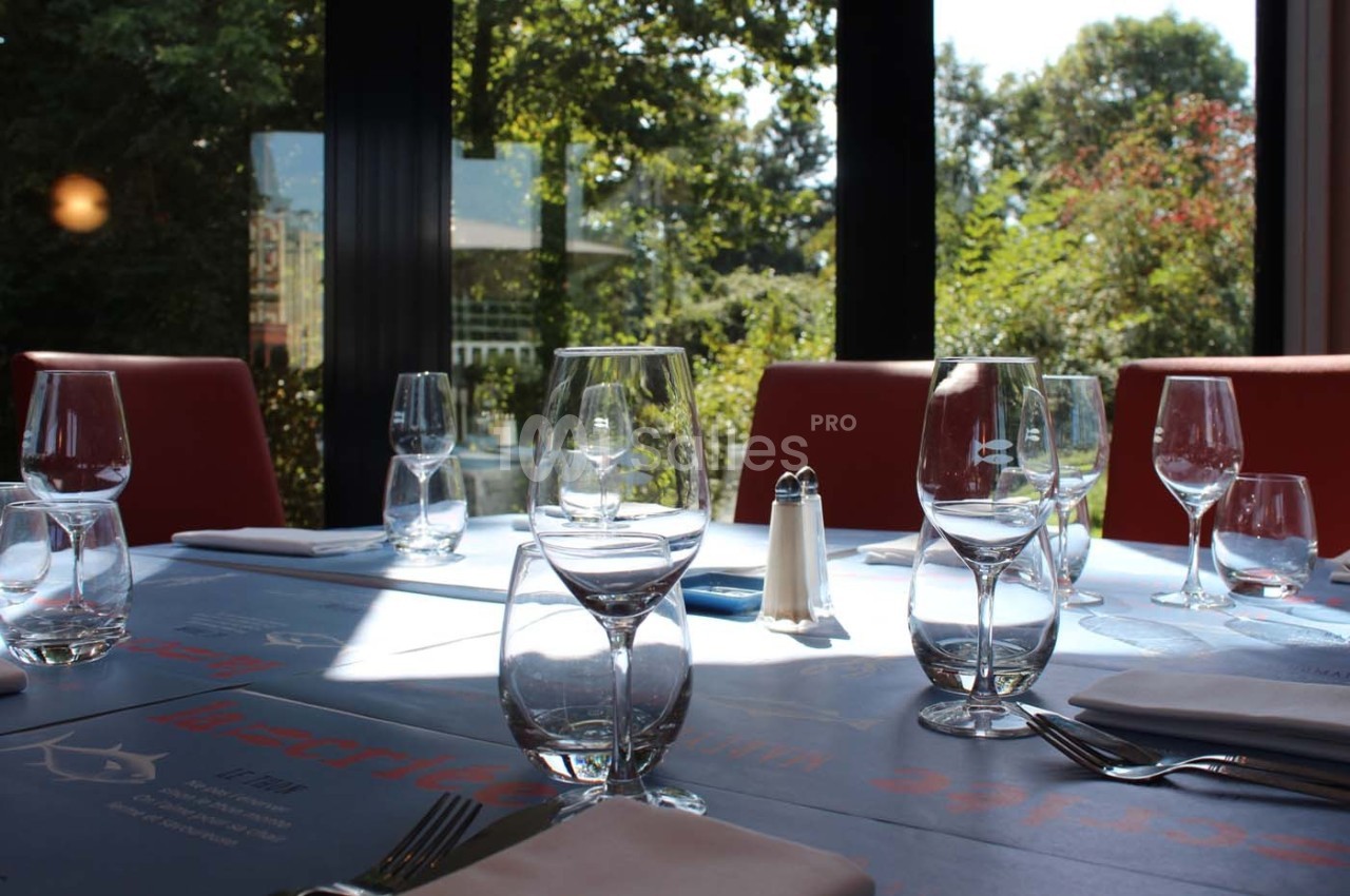 Table dressée avec des verres, couverts et serviettes, près de grandes baies vitrées donnant sur un jardin verdoyant.