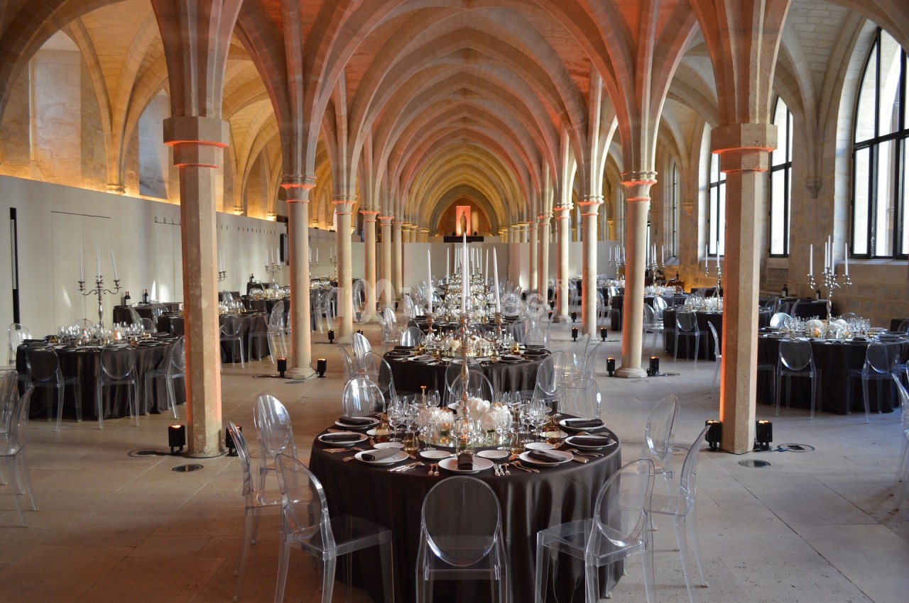 Salle voûtée avec tables rondes dressées pour un événement, chaises transparentes et éclairage chaleureux.