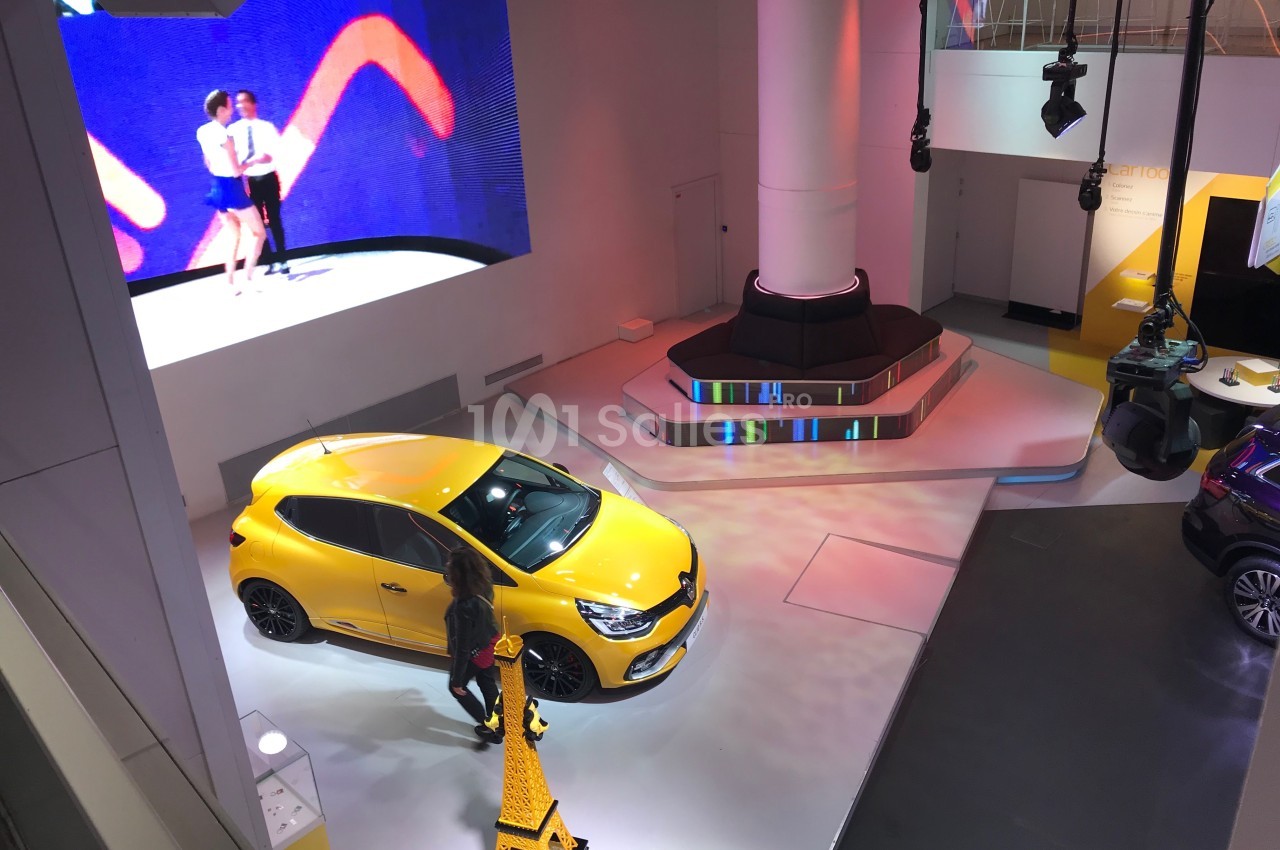 Vue d'une exposition automobile avec une voiture jaune, un écran affichant des danseurs et un éclairage coloré.