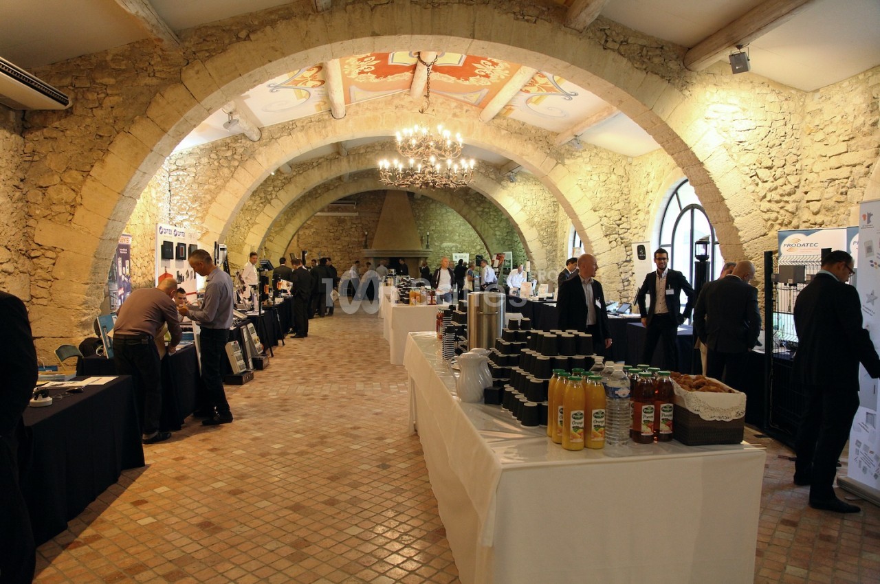 Salle voûtée en pierre accueillant un événement avec des stands, des participants et des tables de boissons.