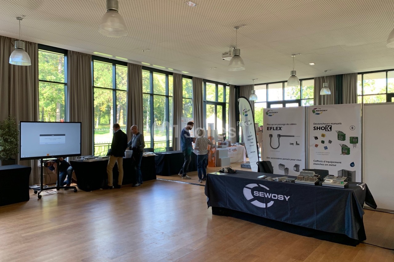 Salle lumineuse avec stands d'exposition, panneaux informatifs et visiteurs discutant près des fenêtres.