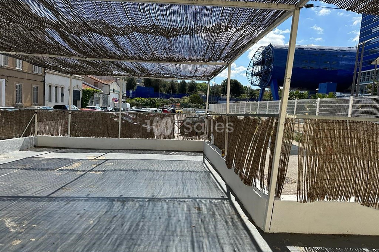 Terrasse ombragée avec des canisses, entourée de bâtiments et d'une structure bleue moderne en arrière-plan.