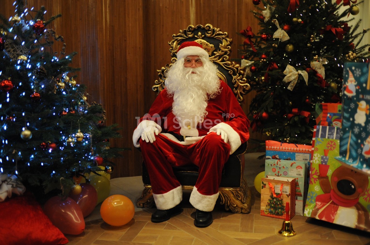Un Père Noël assis sur un fauteuil décoré, entouré de sapins illuminés et de cadeaux colorés.