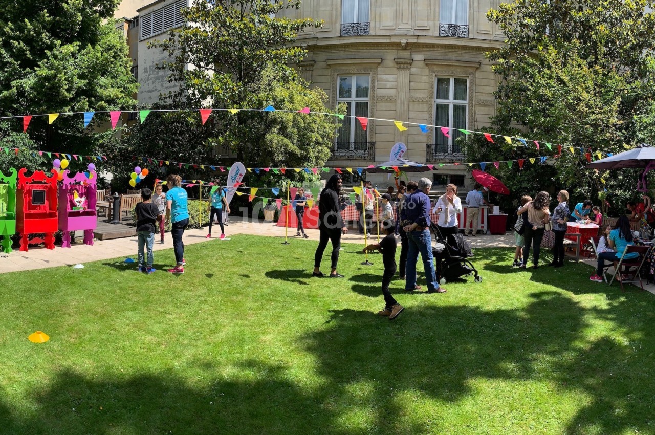 Jardin avec stands colorés, guirlandes de fanions et personnes participant à une fête en plein air.