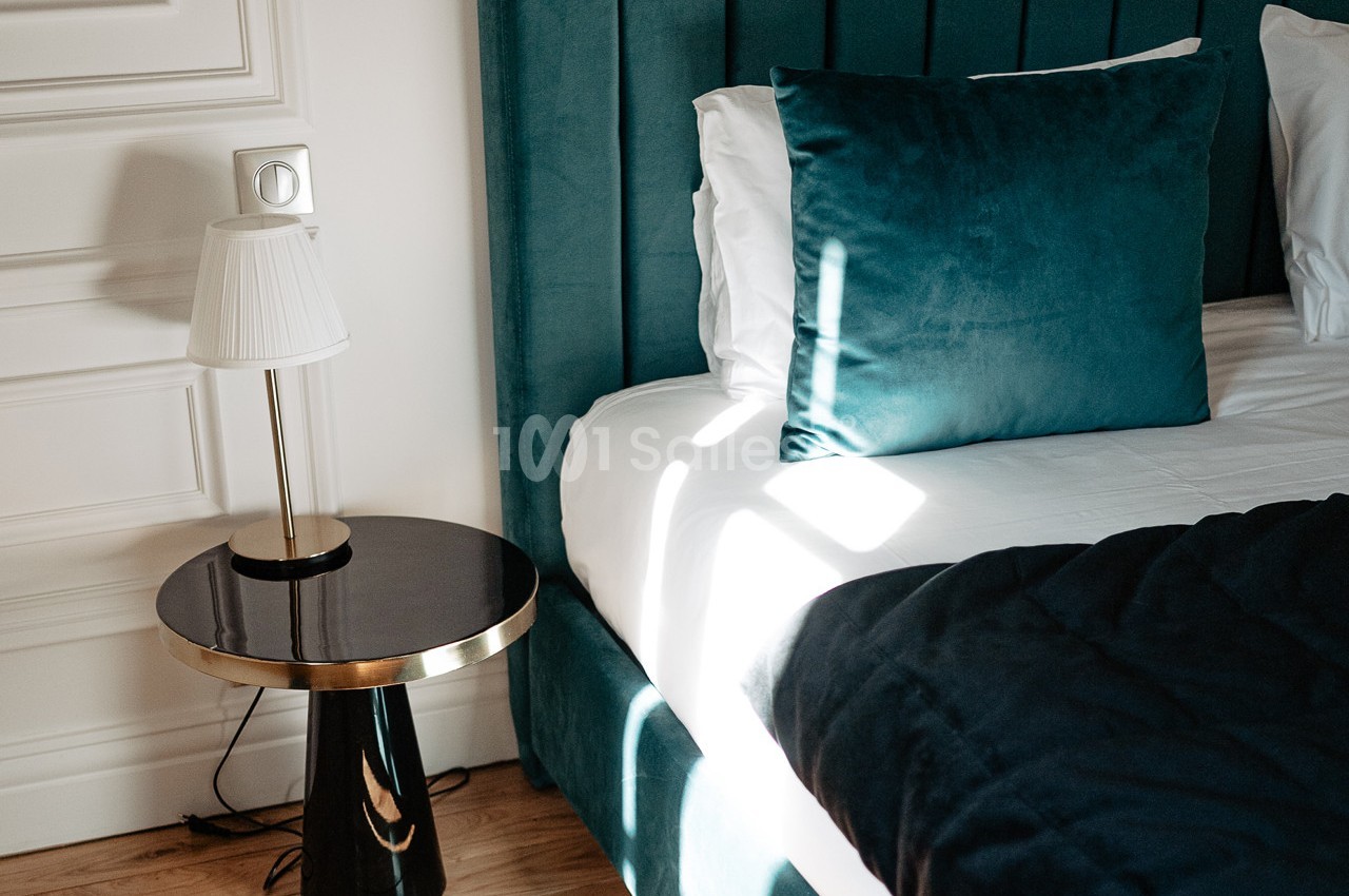 Lit avec tête de lit en velours vert, coussin assorti, table de chevet noire et lampe blanche dans une chambre lumineuse.