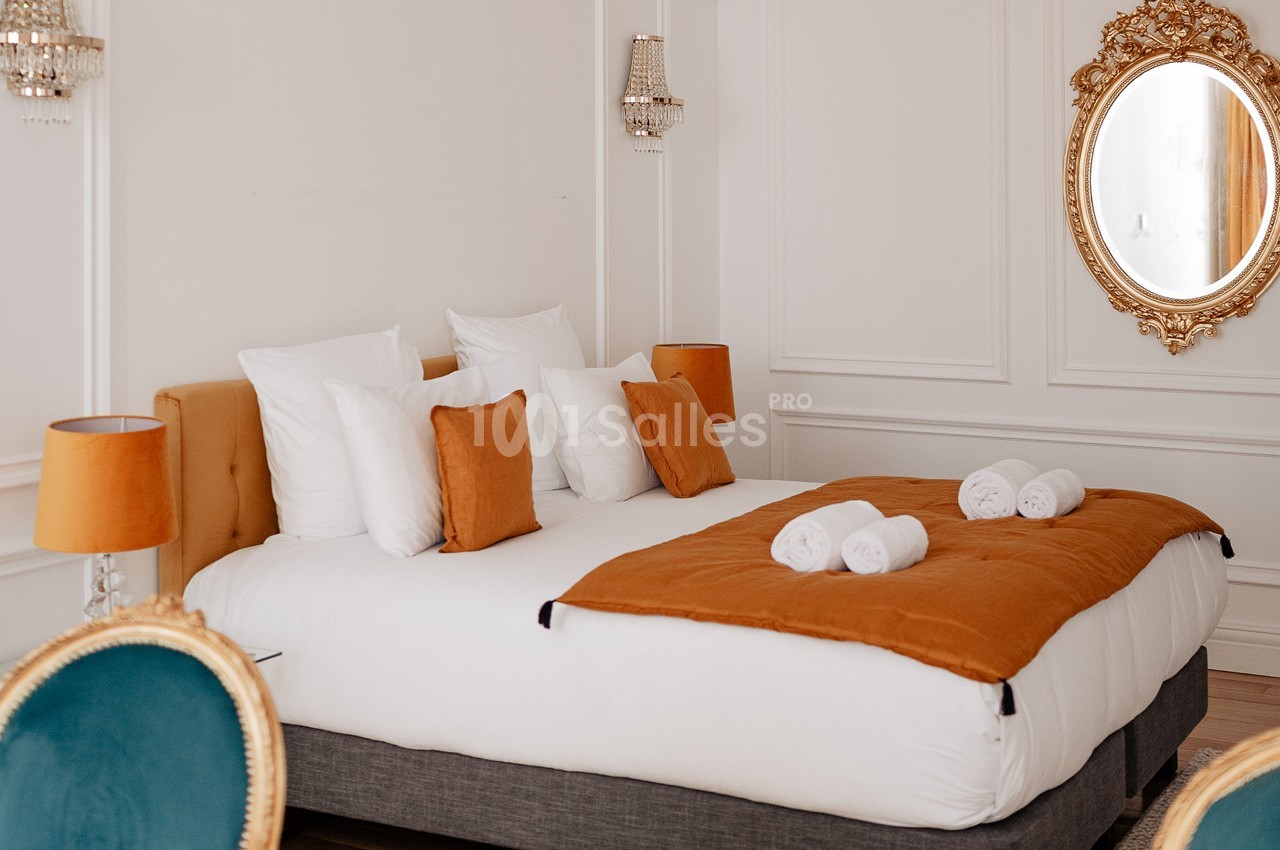 Lit double avec draps blancs, coussins orange, jeté assorti, serviettes pliées, miroir doré et appliques murales.