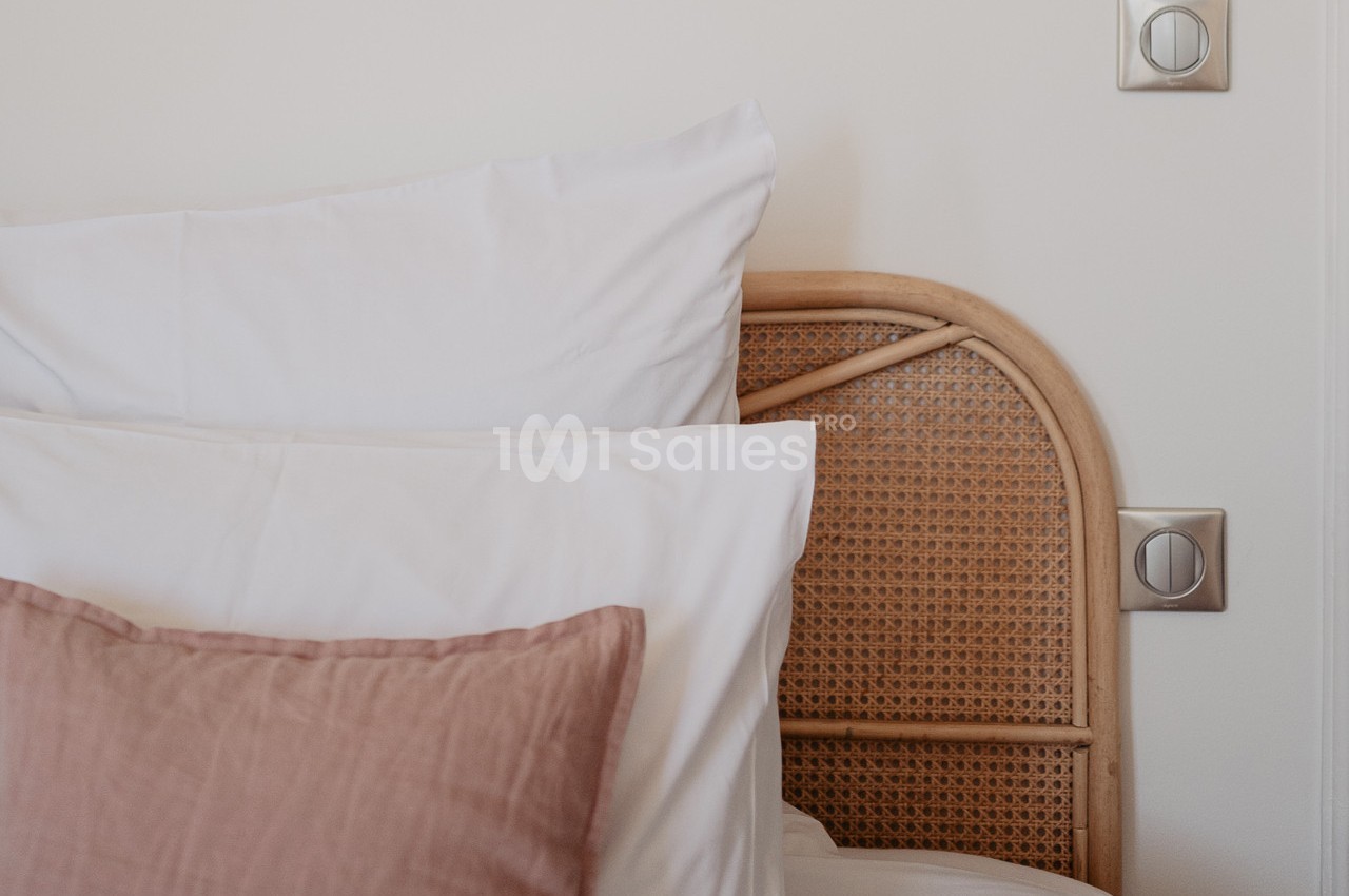 Tête de lit en rotin avec oreillers blancs et un coussin rose, près d'un mur blanc avec deux interrupteurs.