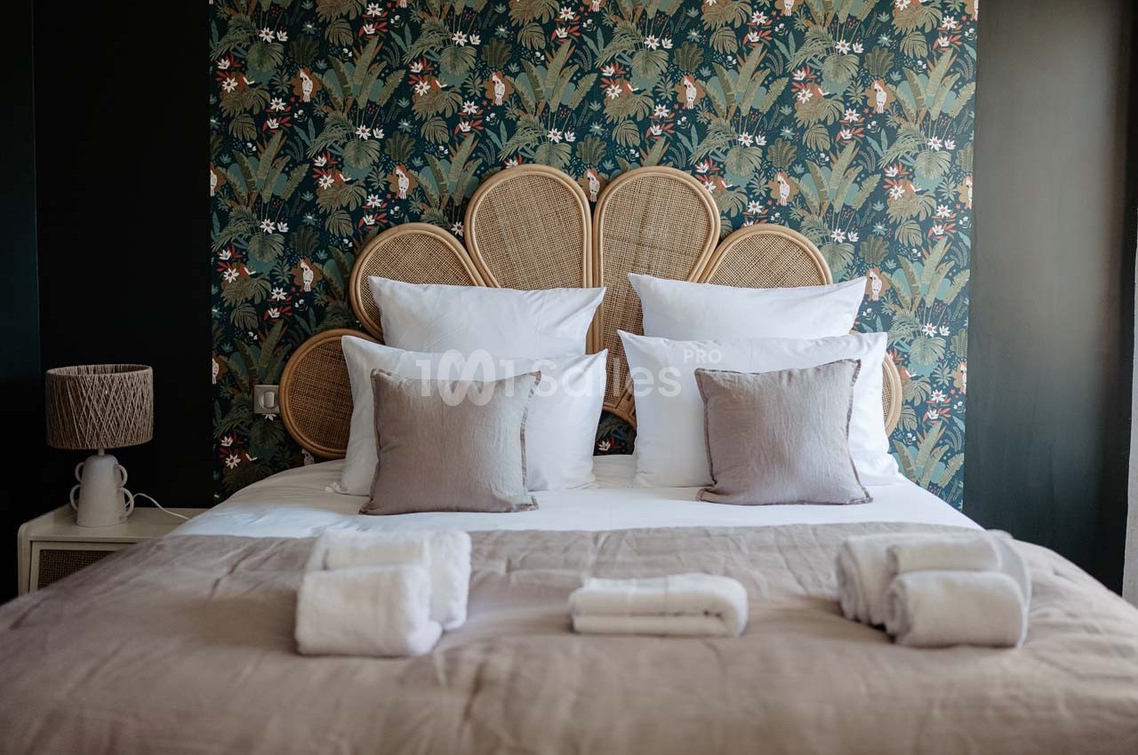 Lit double avec tête de lit en rotin, draps blancs, coussins beiges et fond de papier peint à motifs végétaux.