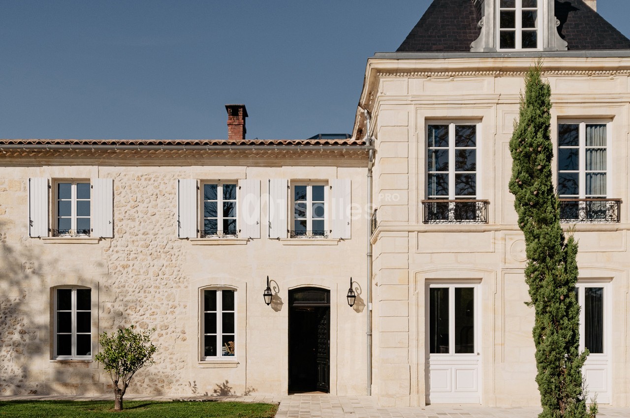 Façade d'une maison en pierre avec volets blancs, toit en ardoise et un cyprès devant l'entrée.
