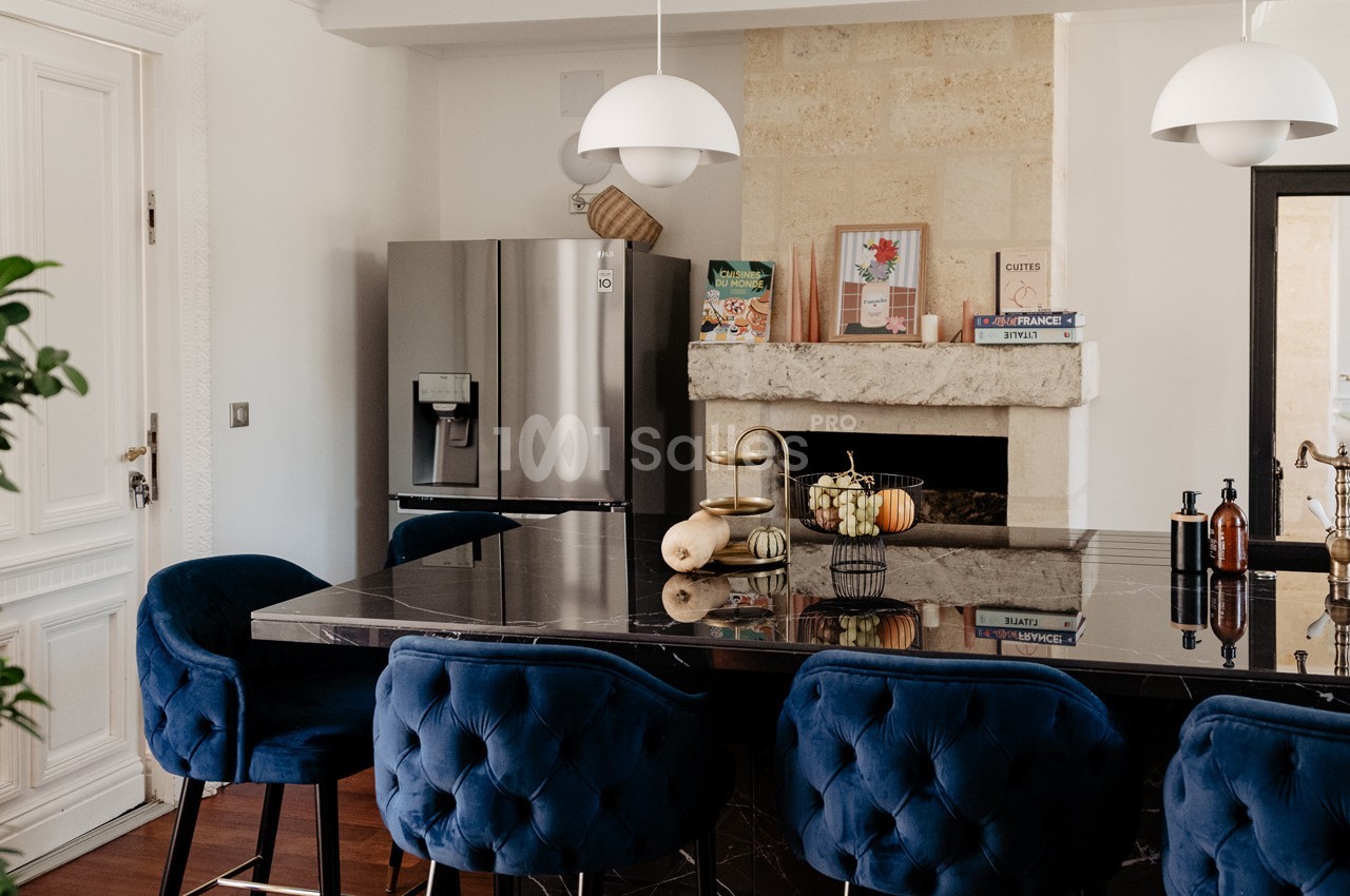 Cuisine moderne avec un îlot central en marbre noir, chaises bleues, réfrigérateur argenté et cheminée décorative.