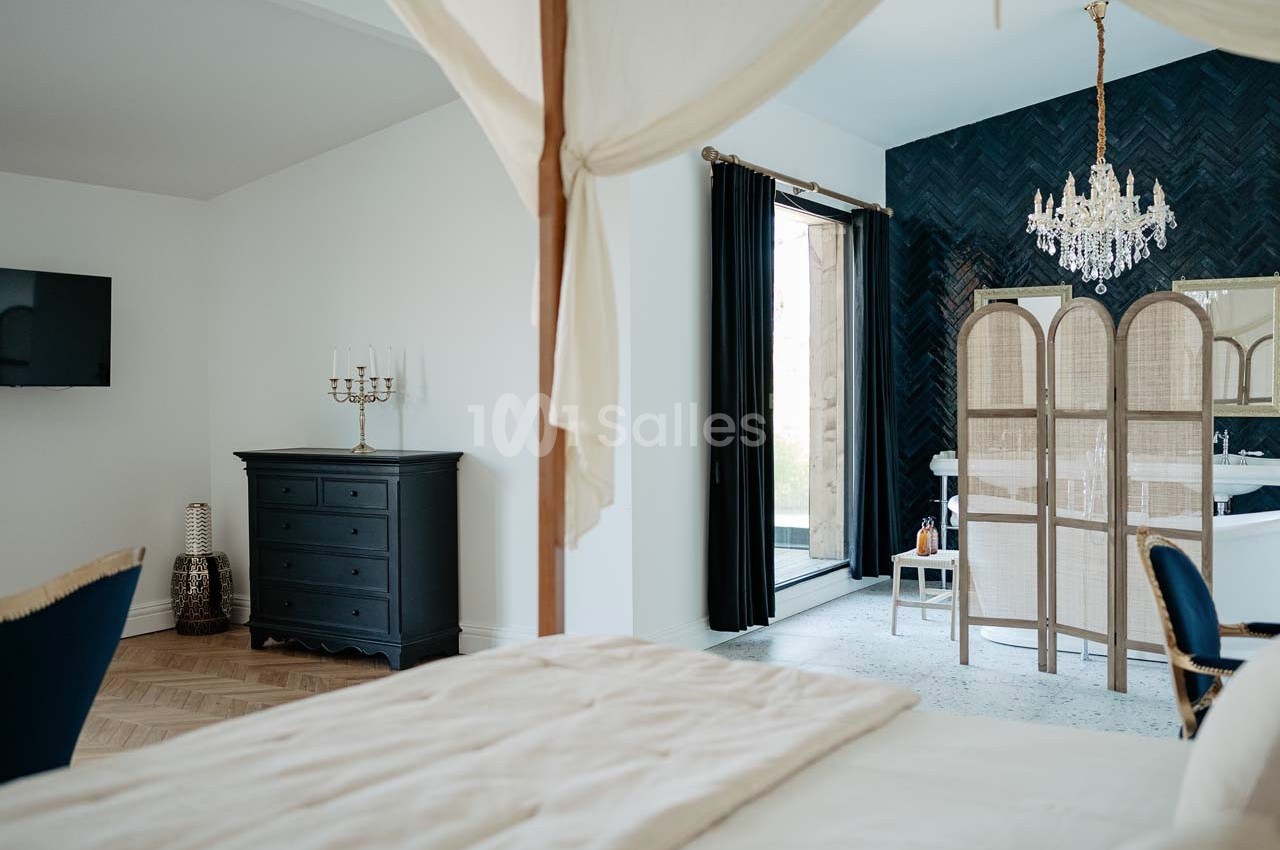 Chambre lumineuse avec lit à baldaquin, commode noire, paravent en rotin, lustre et rideaux sombres.