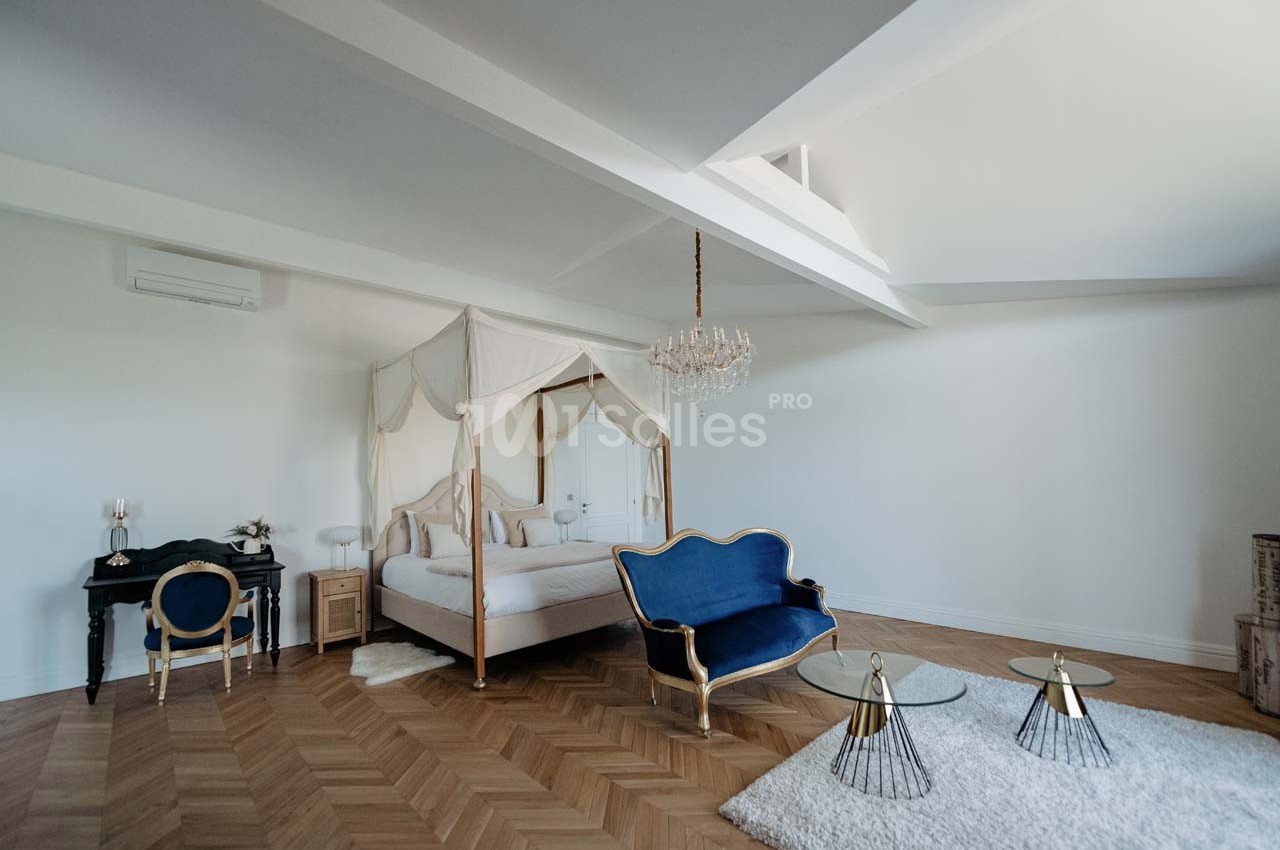 Chambre lumineuse avec lit à baldaquin, canapé bleu, parquet en chevrons et tables basses en verre.