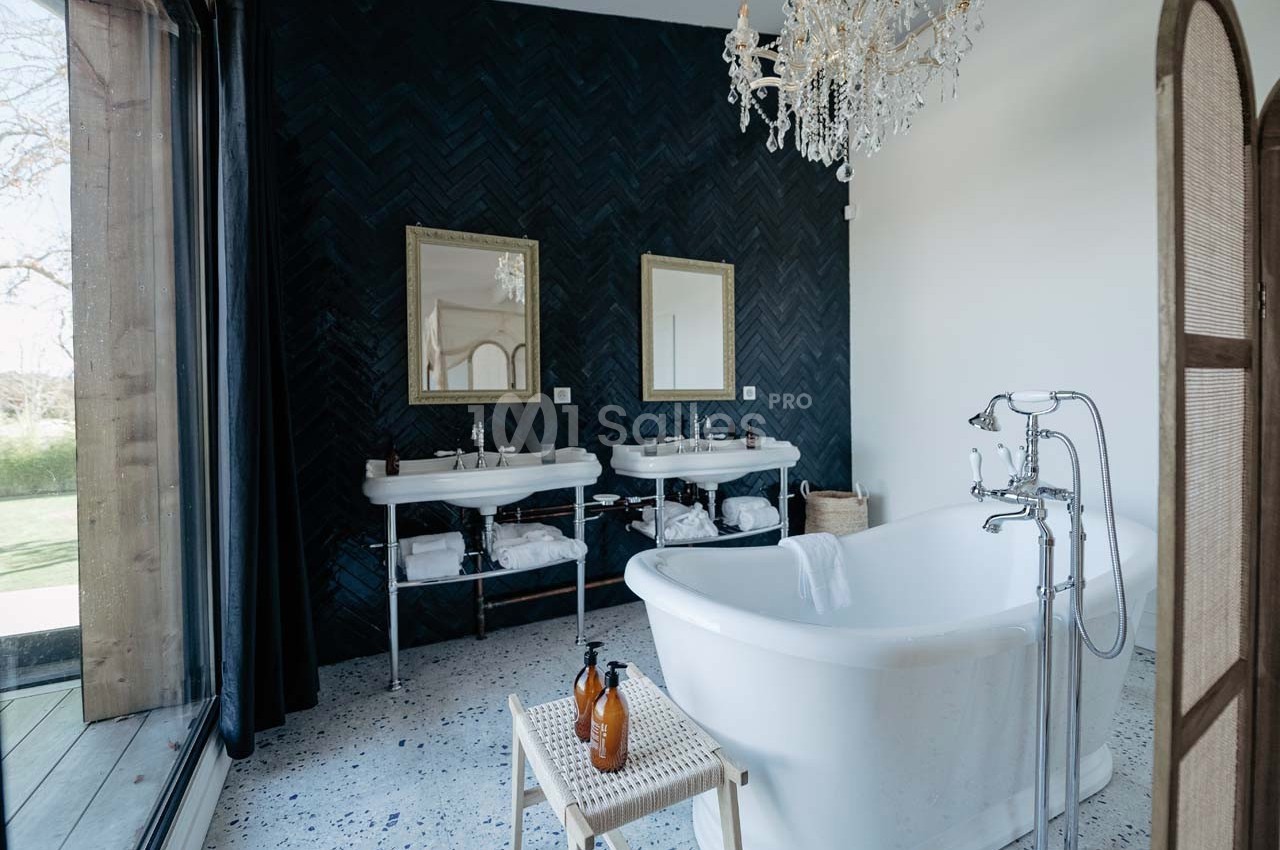 Salle de bain élégante avec baignoire blanche, double vasque, mur sombre en chevrons et lustre en cristal.