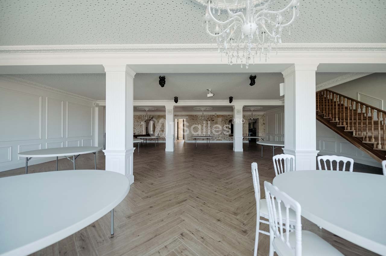 Salle spacieuse avec parquet en chevrons, tables rondes blanches, murs clairs et escalier en bois à droite.