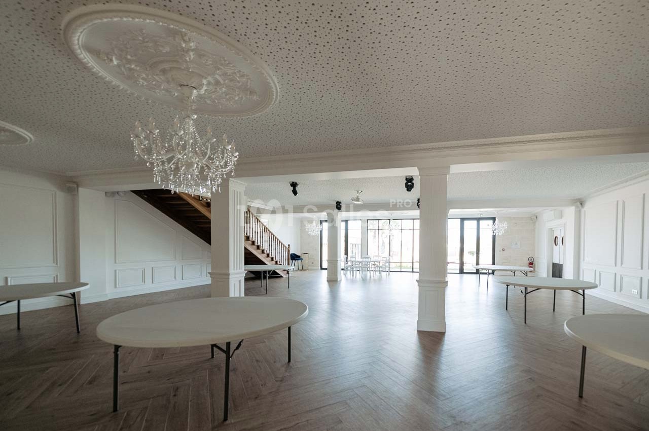 Salle lumineuse avec parquet en bois, tables rondes pliantes, lustres suspendus et escalier en bois sur le côté.