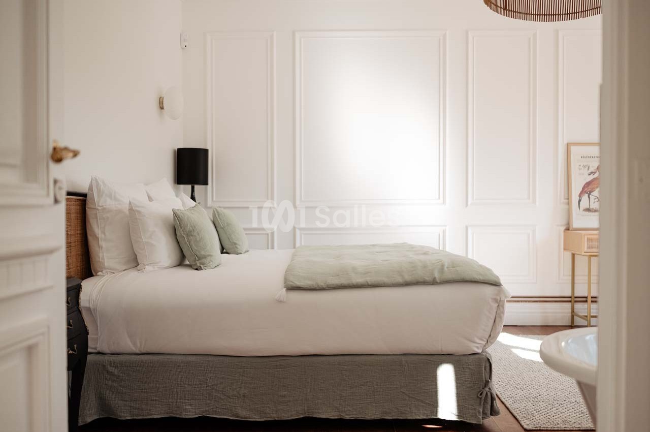 Chambre lumineuse avec lit double, draps blancs, coussins verts, murs blancs moulurés et lampe noire.