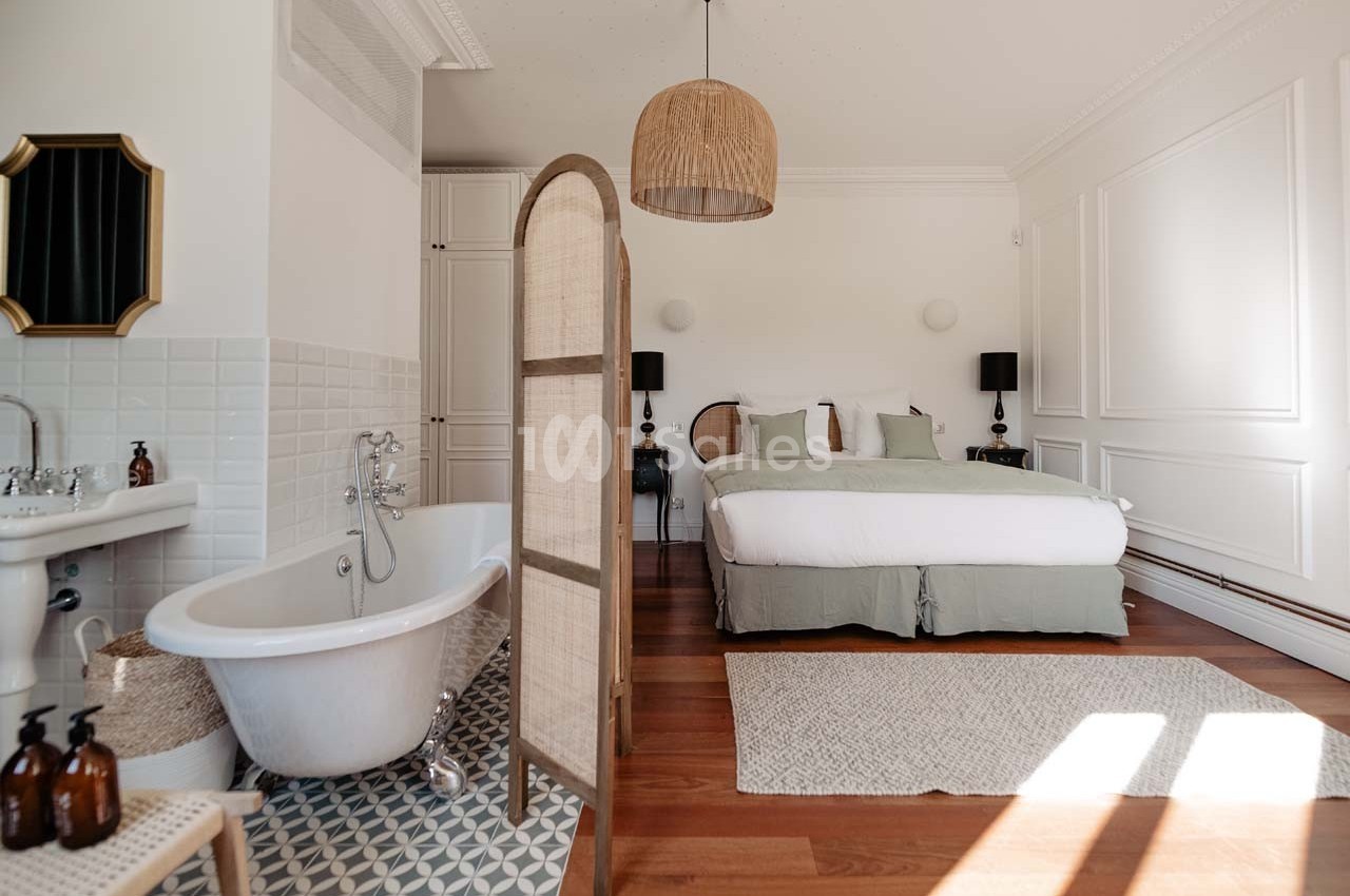 Chambre lumineuse avec lit double, baignoire sur pied, paravent en rotin et décoration élégante dans des tons neutres.