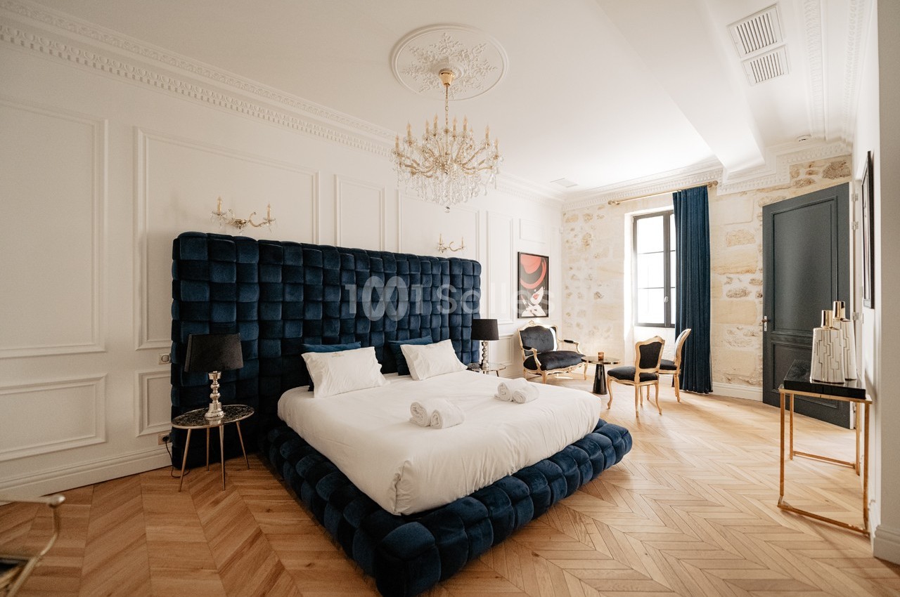 Chambre élégante avec lit double, tête de lit capitonnée bleue, parquet en chevrons et lustre en cristal.