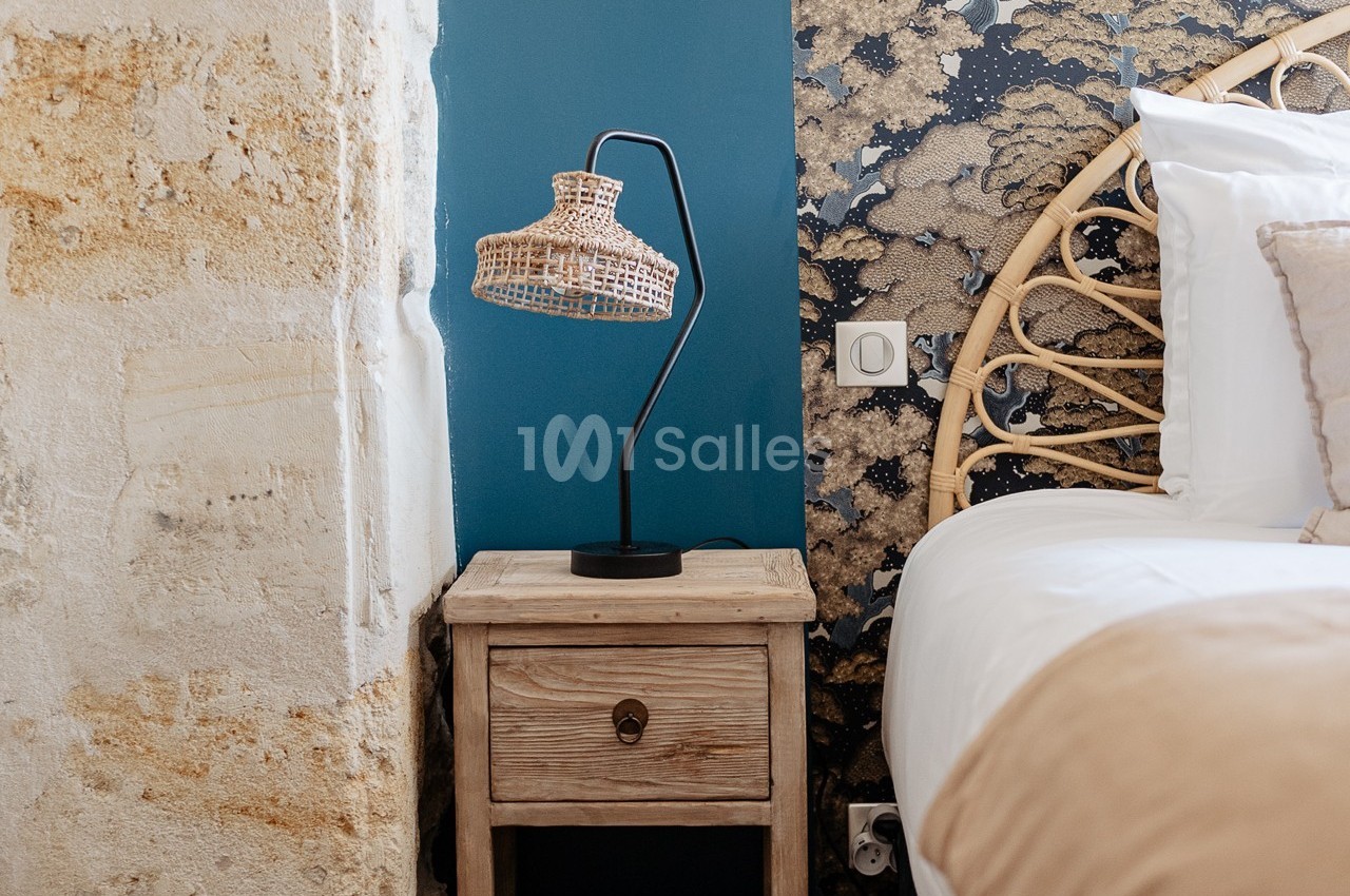 Lampe en osier sur une table de chevet en bois, près d'un lit avec tête de lit en rotin et un mur décoratif.