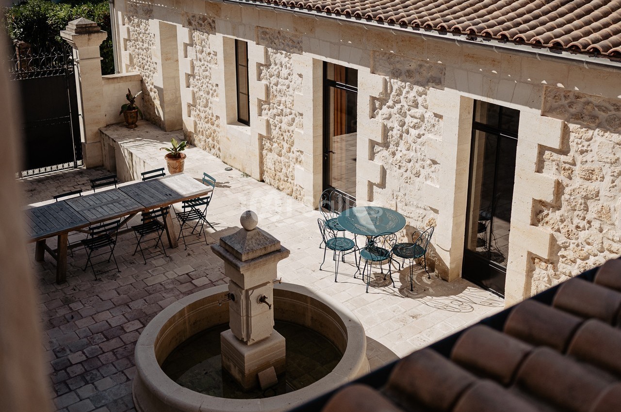 Cour en pierre avec une fontaine centrale, une grande table en bois et des chaises près d'une maison en pierre.