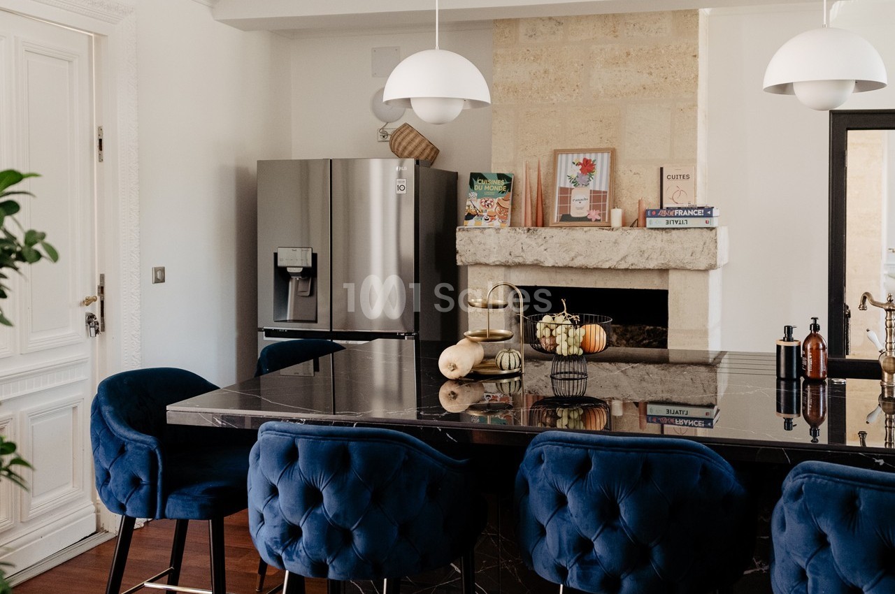 Cuisine moderne avec un îlot central en marbre noir, chaises bleues, réfrigérateur inox et cheminée décorative.
