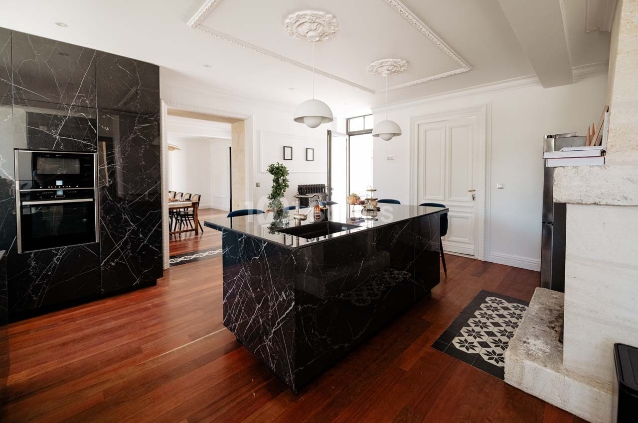Cuisine moderne avec îlot central en marbre noir, parquet en bois, luminaires suspendus et salle à manger en arrière-plan.