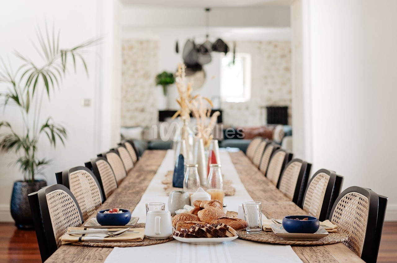 Grande table en bois dressée pour un repas, avec des chaises en rotin, des plats de viennoiseries et des décorations simples.