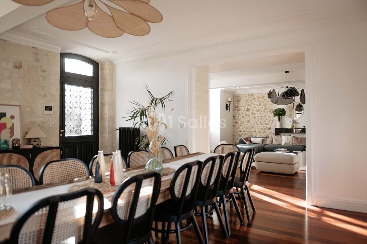 Salle à manger lumineuse avec une grande table en bois, chaises en rotin, et vue sur un salon avec murs en pierre.