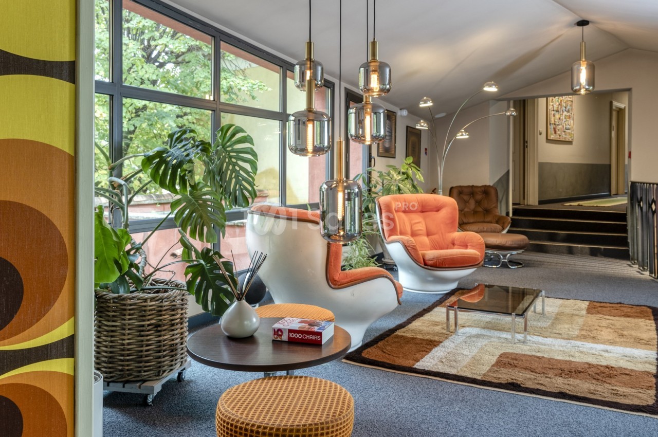 Espace salon rétro avec fauteuils orange, tapis géométrique, plantes vertes et grandes fenêtres donnant sur l'extérieur.