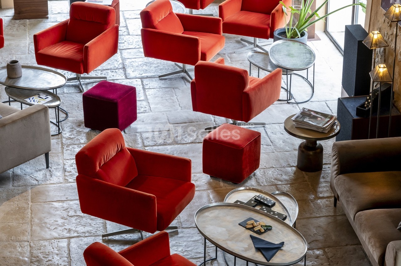Fauteuils rouges et poufs assortis disposés autour de tables basses dans un espace intérieur lumineux au sol en pierre.