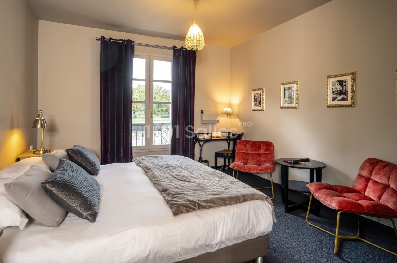Chambre lumineuse avec un lit double, deux fauteuils rouges, un bureau, des cadres au mur et une fenêtre donnant sur l…