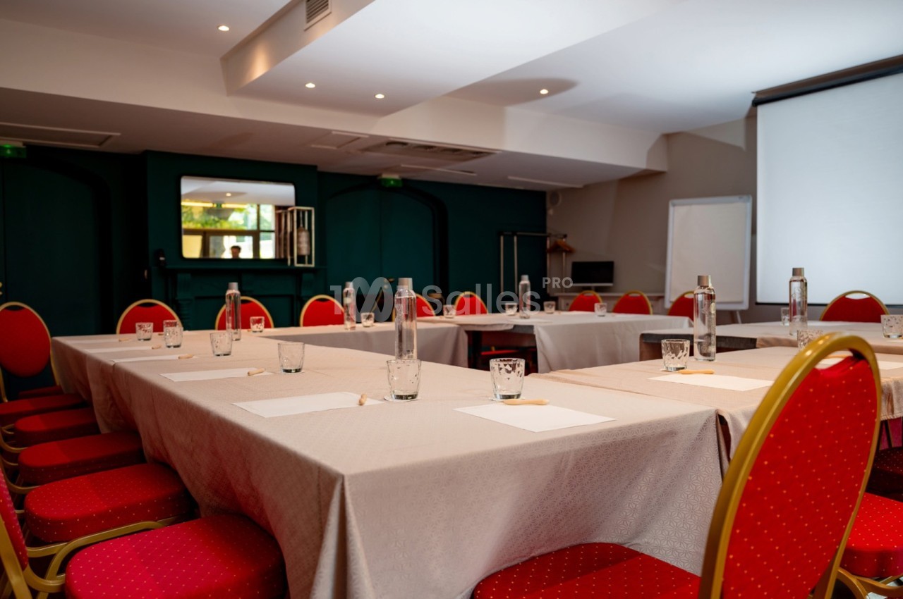 Salle de réunion avec tables en U, chaises rouges, bouteilles d'eau, verres, bloc-notes et écran de projection.