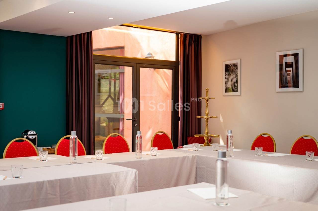 Salle de réunion lumineuse avec tables disposées en U, chaises rouges, bouteilles d'eau et fenêtres donnant sur l'extérieur.