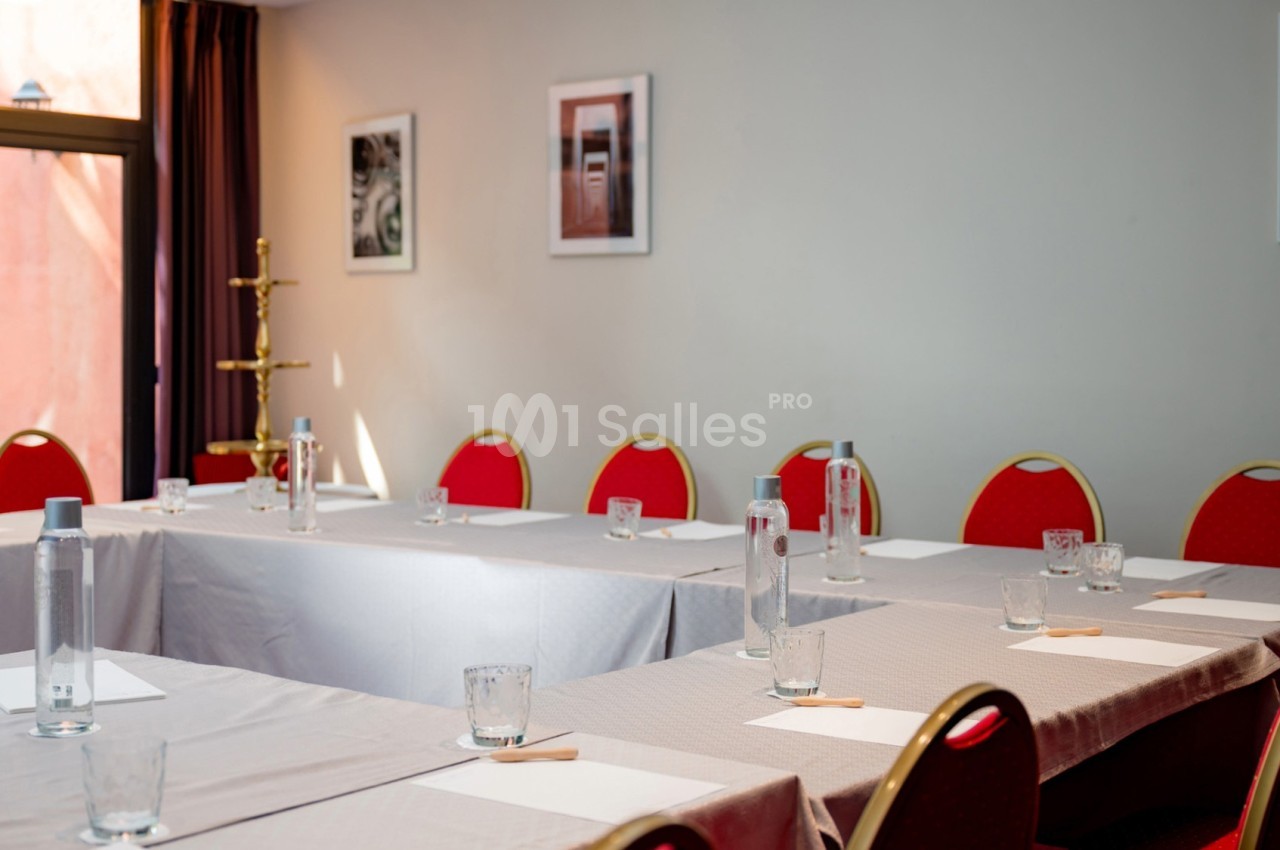 Salle de réunion lumineuse avec des tables en U, chaises rouges et bouteilles d'eau disposées sur les tables.